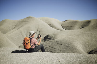 Backpacker photographing through mobile phone while sitting on sand at desert against clear sky - CAVF53142