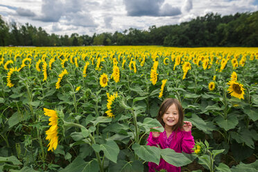 Porträt eines niedlichen glücklichen Mädchens, das auf einer Sonnenblumenfarm vor einem bewölkten Himmel steht - CAVF53150