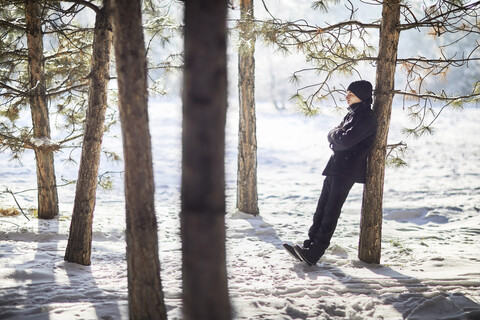 Junger Mann in voller Länge in schwarzer warmer Kleidung, der sich im Winter im Wald an einen Baumstamm lehnt, lizenzfreies Stockfoto