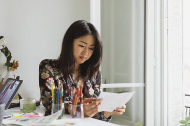 Illustratorin sitzt am Schreibtisch im Atelier und betrachtet eine Zeichnung - AFVF01936