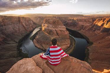 USA, Arizona, Colorado River, Horseshoe Bend, junger Mann sitzt auf Aussichtspunkt mit amerikanischer Flagge - KKAF02844