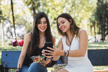 Freundinnen sitzen auf einer Parkbank und schauen auf ihr Smartphone - KKAF02871