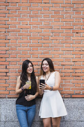 Girl frinds standing in front of a brick wall, looking at smartphone - KKAF02876