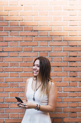 Lachende junge Frau vor einer Backsteinmauer mit Smartphone - KKAF02877