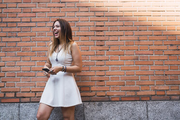 Laughing young woman in front of a brick wall using smartphone - KKAF02878