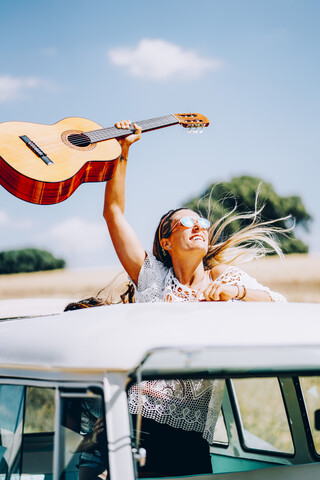 Happy young woman holding her guitar aloft, standing in her van on a road trip stock photo