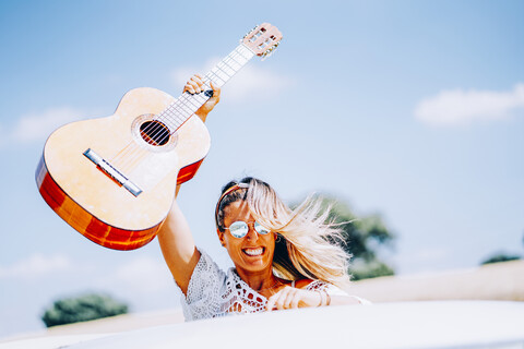 Glückliche junge Frau, die ihre Gitarre in die Höhe hält, während sie in ihrem Van auf einer Autoreise steht, lizenzfreies Stockfoto