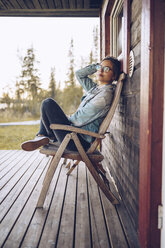 Schweden, Lappland, Porträt einer jungen Frau, die auf einem Stuhl auf einer Veranda sitzt und sich entspannt - RSGF00025