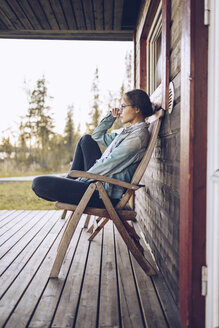Schweden, Lappland, nachdenkliche junge Frau sitzt auf einem Stuhl auf der Veranda und schaut in die Ferne - RSGF00026