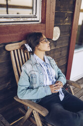 Sweden, Lapland, pensive young woman sitting on chair on veranda looking at distance - RSGF00027