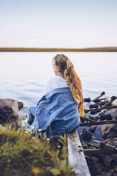 Schweden, Lappland, Rückenansicht einer jungen Frau, die am Wasser sitzt und in die Ferne schaut - RSGF00029