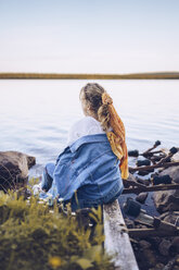 Sweden, Lapland, back view of young woman sitting at water's edge looking at distance - RSGF00029