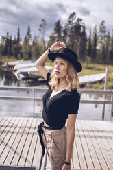 Sweden, Lapland, portrait of fashionable blond woman wearing black hat standing on jetty - RSGF00038