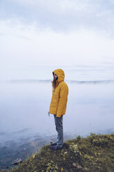 Sweden, Lapland, man with full beard wearing yellow windbreaker standing at water's edge looking at distance - RSGF00045