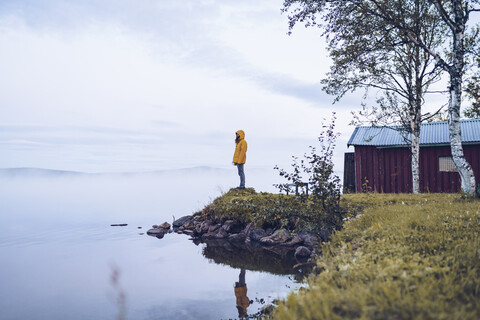 Schweden, Lappland, Mann mit Windjacke steht am Wasser und schaut in die Ferne, lizenzfreies Stockfoto