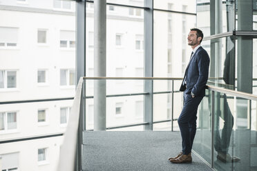 Successful businessman standing in office building, looking out of window, daydreaming - UUF15794