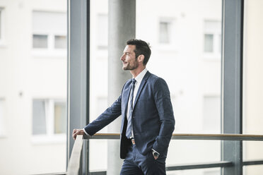 Successful businessman standing in office building, looking out of window, daydreaming - UUF15795