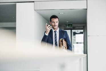 Businessman with briefcase walking in office building, using smartphone - UUF15797