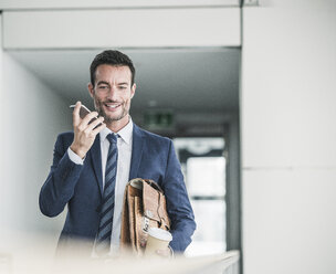 Businessman with briefcase walking in office building, using smartphone - UUF15798