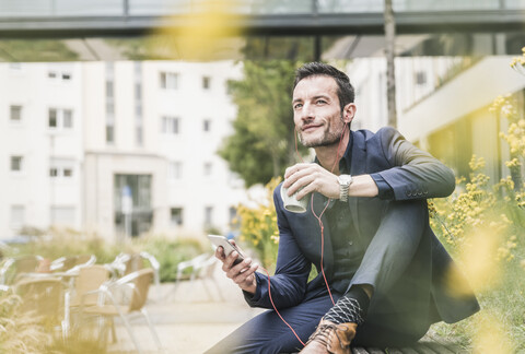 Geschäftsmann sitzt draußen, macht eine Pause, hört Musik von seinem Smartphone und trinkt Kaffee, lizenzfreies Stockfoto