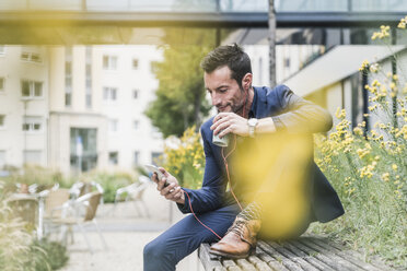 Businessman sitting outside, taking a break, listening music from his smartphone, drinking coffee - UUF15815