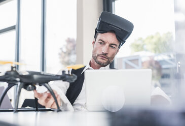 Businessman sitting in office working on a drone, using VR glasses - UUF15854