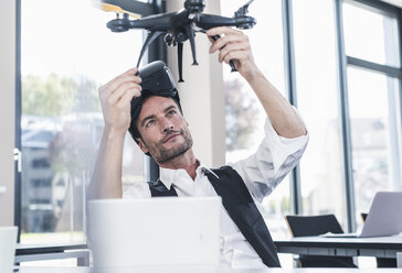 Businessman sitting in office working on a drone, using VR glasses - UUF15855