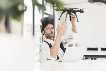 Businessman sitting in office working on a drone, using VR glasses - UUF15856