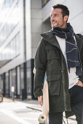 Mature man, wearing coat and scarf, walking in the city, carrying longboard - UUF15861