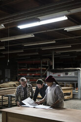 Drei gemischtrassige Tischler, die nach Feierabend in einer großen Holzverarbeitungsfabrik an einem Laptop-Computer arbeiten. - MINF09115