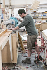 Caucasian male carpenter using a radial saw to cut a piece of wood in a large woodworking factory. - MINF09135