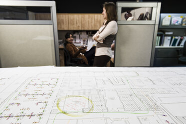 Closeup of plans on a table with two people working in the background. - MINF09148