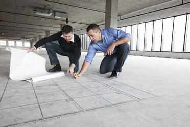 An architect and a business owner crouching over architectural plans and drawing on a concrete floor, planning a new office space in an empty raw built space. - MINF09156