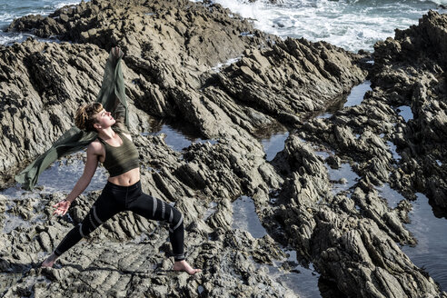 Junge Frau mit braunem Haar und Dreadlocks in Sportkleidung steht am felsigen Ufer am Meer und macht Tai Chi. - MINF09162