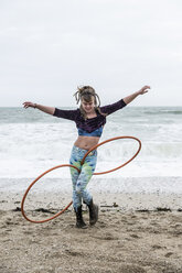 Junge Frau mit braunen Haaren und Dreadlocks steht an einem Sandstrand am Meer und balanciert zwei Hula-Hoop-Reifen. - MINF09166