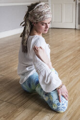 Junge Frau mit Kopftuch und weißer Bluse sitzt in Yoga-Pose auf dem Boden. - MINF09169