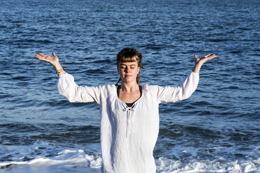 Junge Frau mit braunem Haar und weißer Bluse steht am Strand am Meer und macht Tai Chi. - MINF09170