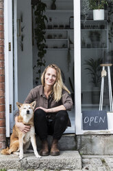 Lächelnde Inhaberin eines Pflanzenladens, die auf den Stufen vor ihrem Geschäft sitzt, ein Hund sitzt neben ihr. - MINF09173