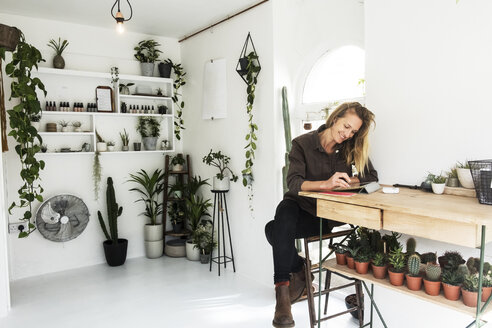 Female owner of plant shop sitting at table, working on digital tablet, a selection of plants on wooden shelves. - MINF09177