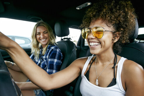 Porträt von zwei Frauen mit langen blonden und braunen lockigen Haaren, die im Auto sitzen, Sonnenbrillen tragen und lächeln. - MINF09204