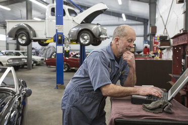 A senior Caucasian male car mechanic working on his laptop computer in his classic car repair shop. - MINF09233