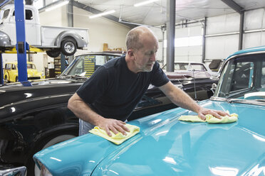 A Caucasian senior male car mechanic applying wax to the hood of an old sedan in his classic car repair shop. - MINF09235