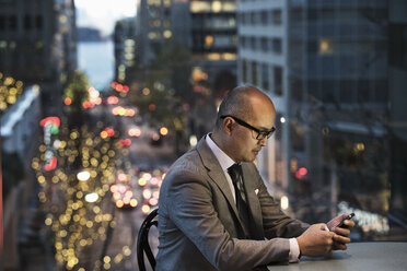 Seitenansicht eines asiatischen Geschäftsmannes, der an einem Tisch sitzt und an seinem Telefon arbeitet, vor einem Fenster mit Blick auf eine Straße in der Abenddämmerung. - MINF09272
