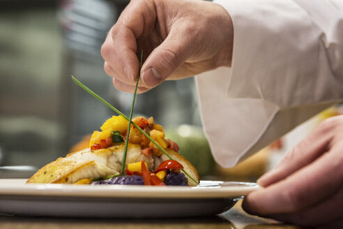 A closeup of the hands of a chef putting the final dressing on a plate of fish in a commercial kitchen, - MINF09298