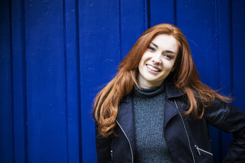Portrait of smiling young woman with long red hair in front of bright blue door. - MINF09326