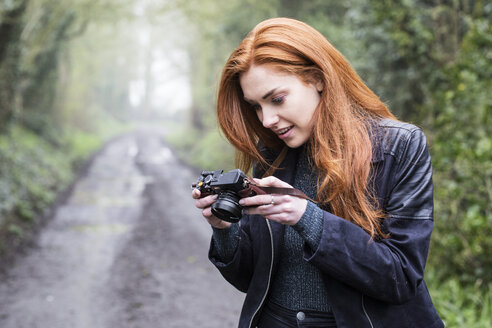 Lächelnde junge Frau mit langen roten Haaren, die einen Waldweg entlangläuft und mit einer alten Kamera Fotos macht. - MINF09328