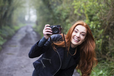 Lächelnde junge Frau mit langen roten Haaren, die einen Waldweg entlangläuft und mit einer alten Kamera Fotos macht. - MINF09330