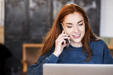 Lächelnde junge Frau mit langen roten Haaren sitzt am Tisch, arbeitet am Laptop und benutzt ein Mobiltelefon. - MINF09331