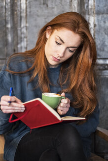 Junge Frau mit langen roten Haaren sitzt an einem Tisch, in der Hand ein Notizbuch und eine Tasse Kaffee. - MINF09333