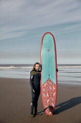 Porträt eines Mannes, der ein dekoriertes Surfbrett an einem Sandstrand hält - MINF09390